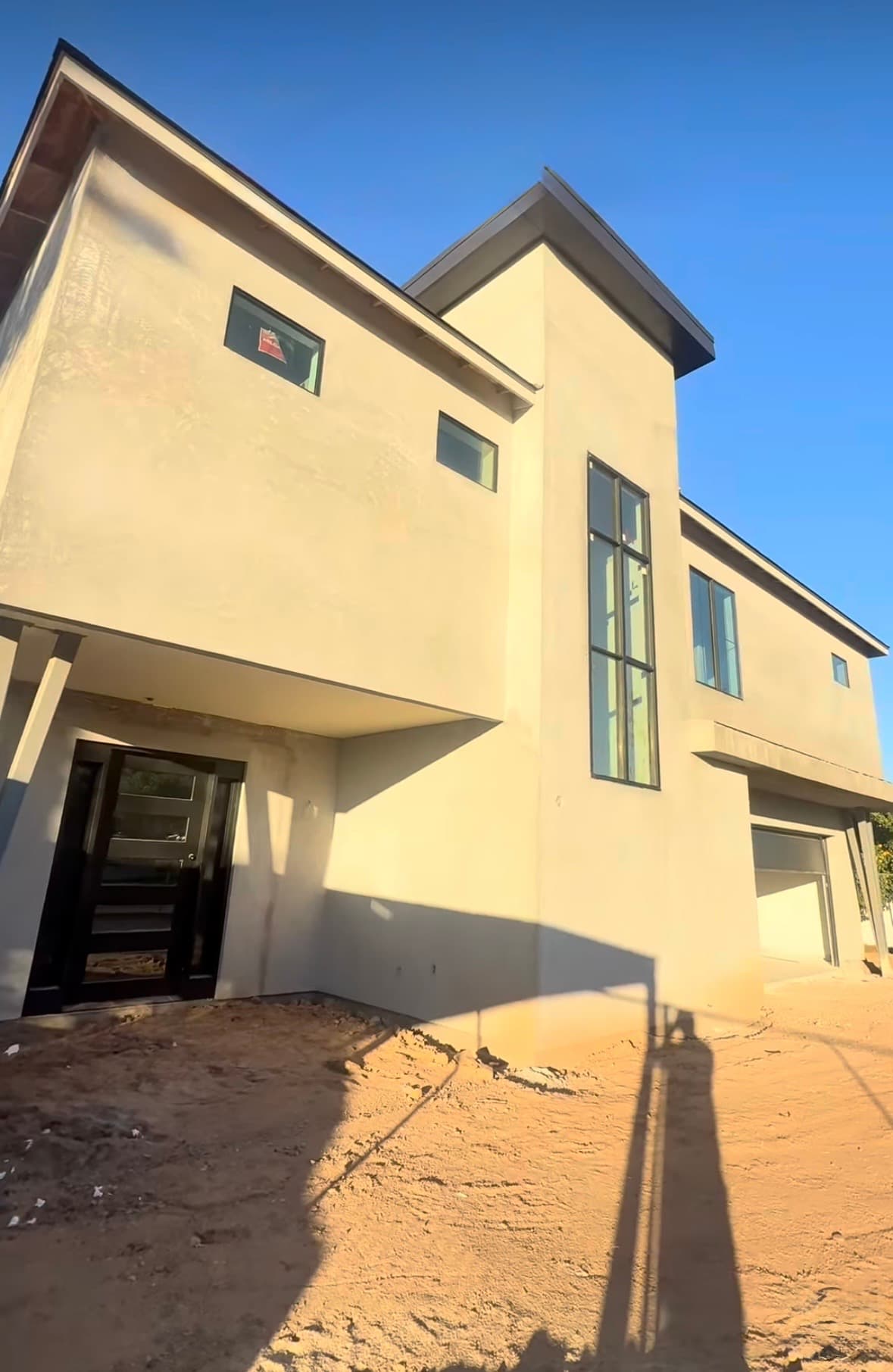 Contemporary light-colored house under construction, "For Sale" sign in window, bare dirt ground.