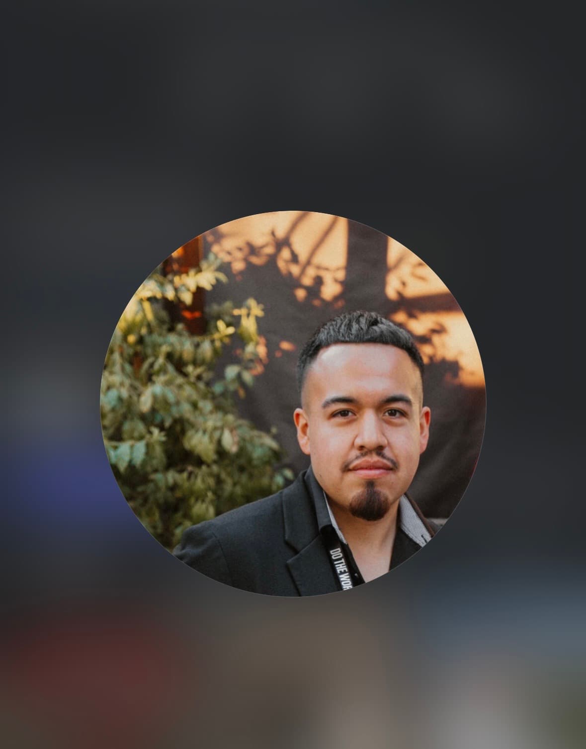 Man with short dark hair, mustache, and goatee wearing a black blazer against leafy greenery.