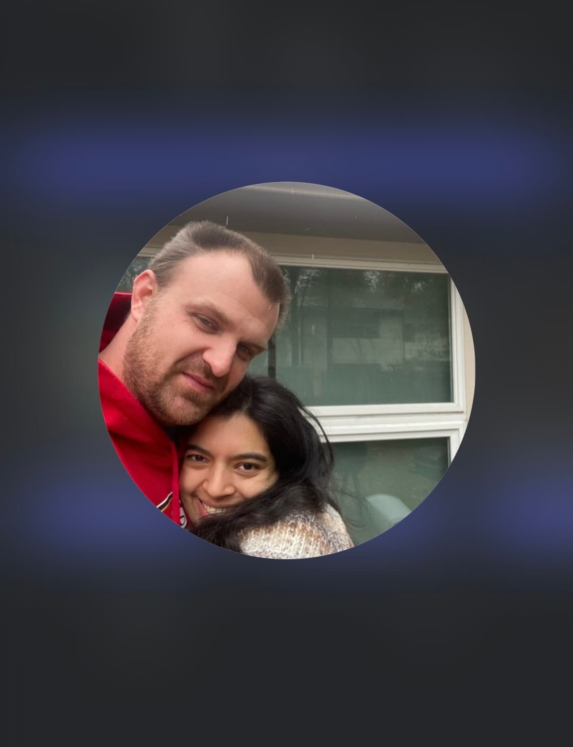 Bearded man in a red hoodie and a smiling woman pose together outside a window.
