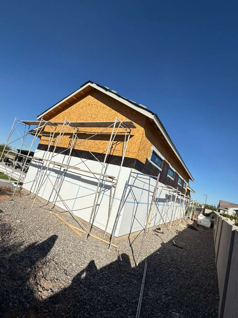 House frame with plywood sheathing, white exterior panels, and scaffolding under blue sky.