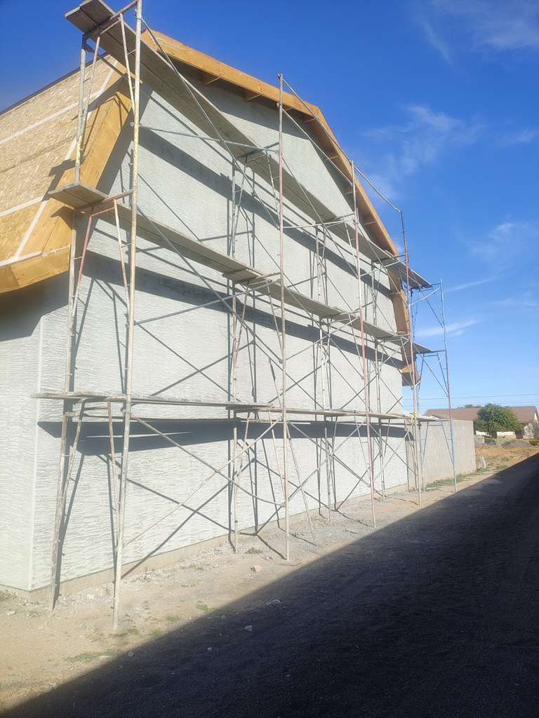 Scaffolding covers a light-colored house under construction with exposed roof framing and blue sky.