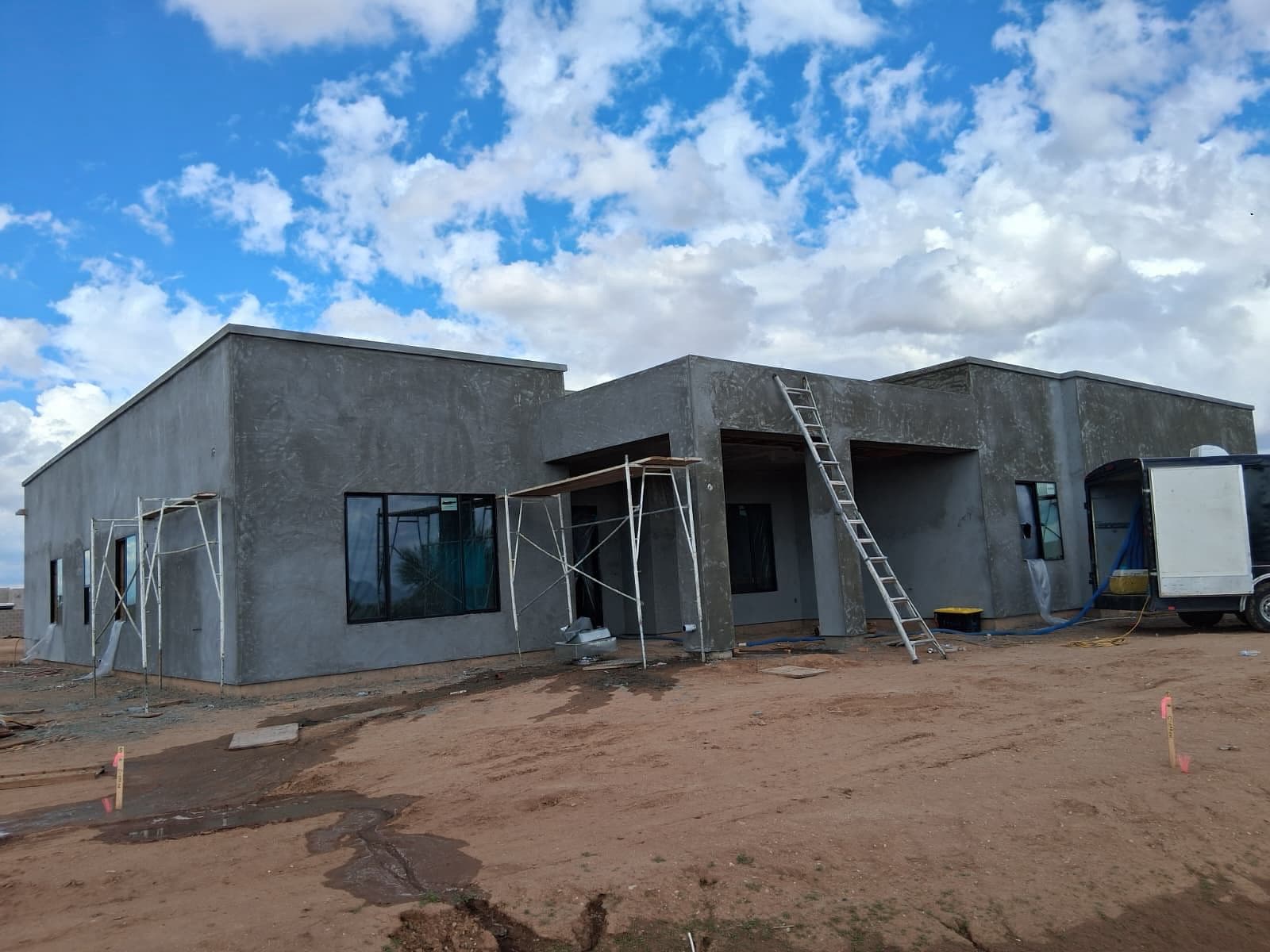 Contemporary gray stucco house under construction with scaffolding, ladder, and dirt yard.