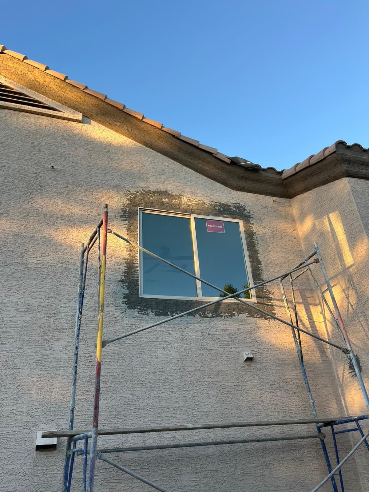 House wall with new Milgard window, wet stucco patch, scaffolding, and blue sky.